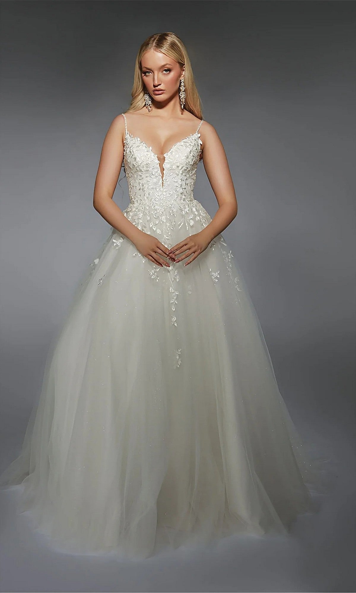 A woman stands against a gray background wearing the Long White Dress: Alyce 7127, featuring a fitted bodice, lace and floral embroidery details, thin straps, and a full tulle skirt.
